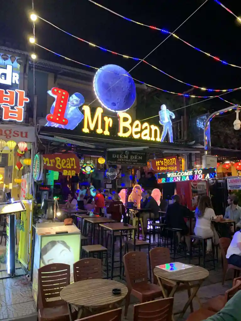 A nighttime photo of a busy outdoor bar called 'Mr Bean Bar' in Hoi An, Vietnam, featuring neon signs, string lights, and…