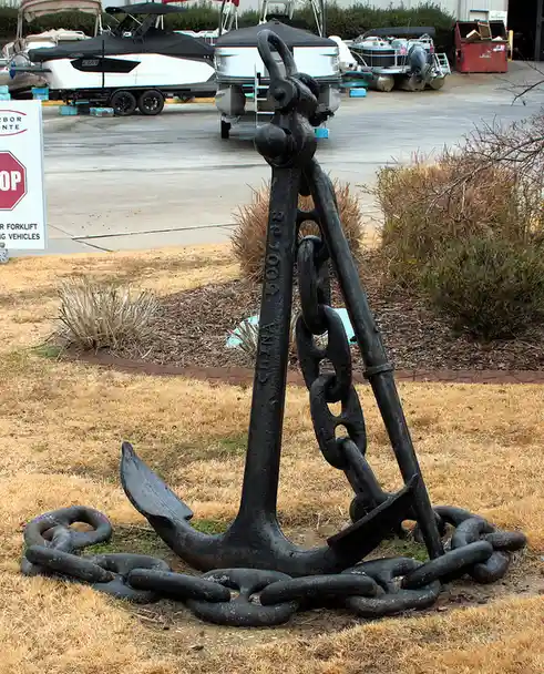 A large, black, rusty anchor with a heavy chain lies on dry, brown grass in front of a boat storage facility. Several boats…