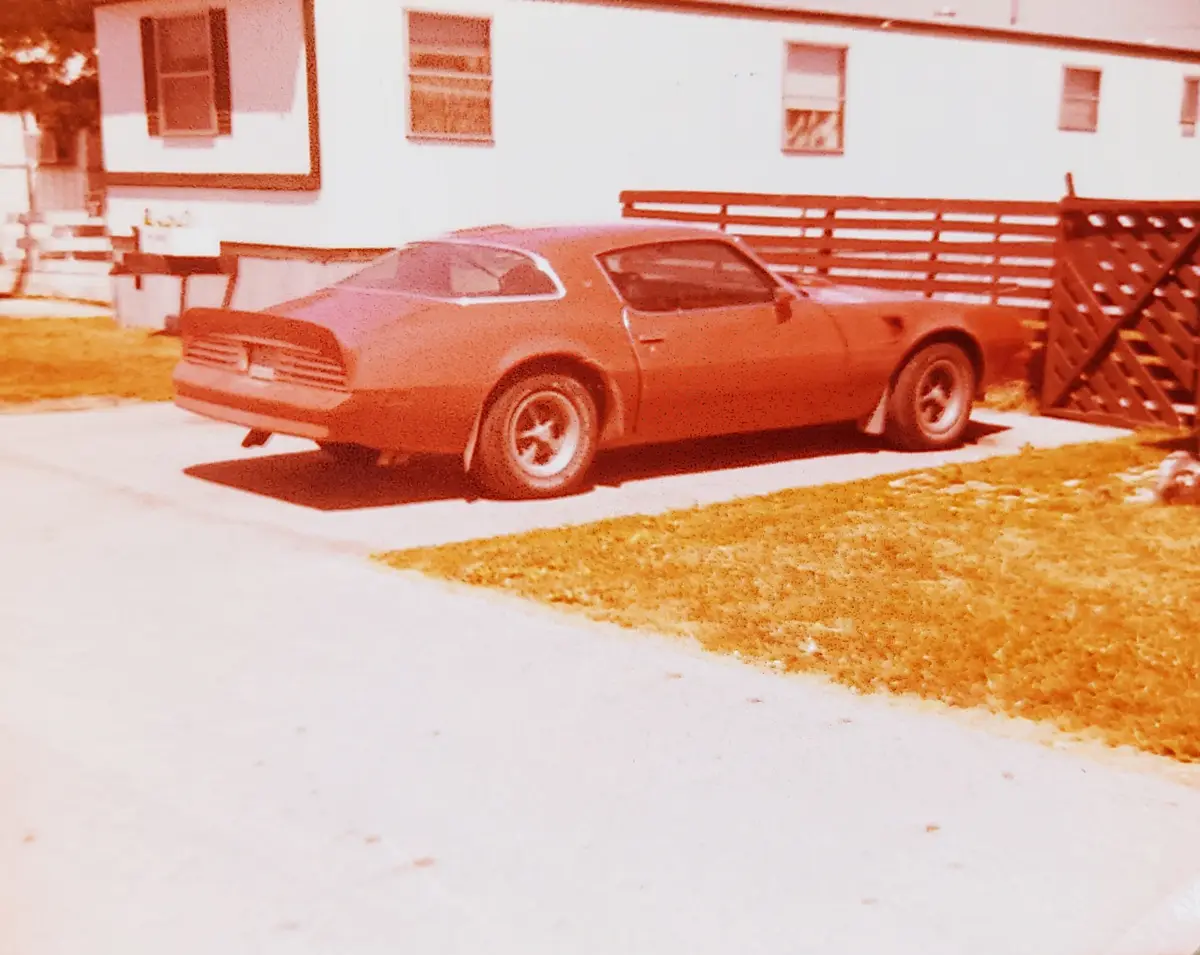 A vintage red muscle car, likely a Pontiac Firebird, is parked on a concrete driveway in front of a white mobile home with a…