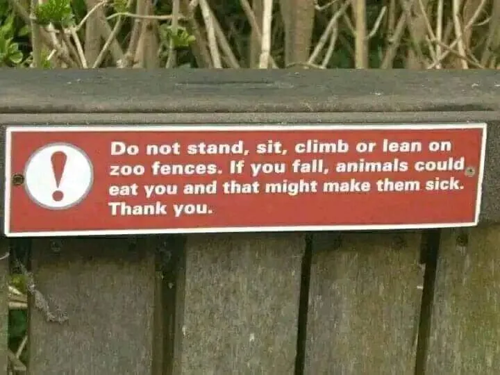 A red warning sign with white text and an exclamation mark icon is mounted on a wooden zoo fence. The sign reads: 'Do not…