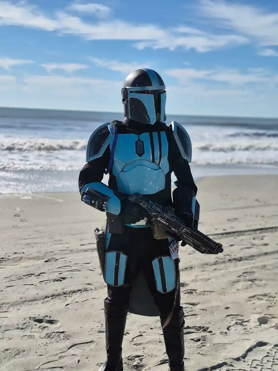 A person in a blue and black Mandalorian-style cosplay armor stands on a sandy beach holding a blaster rifle, with ocean…