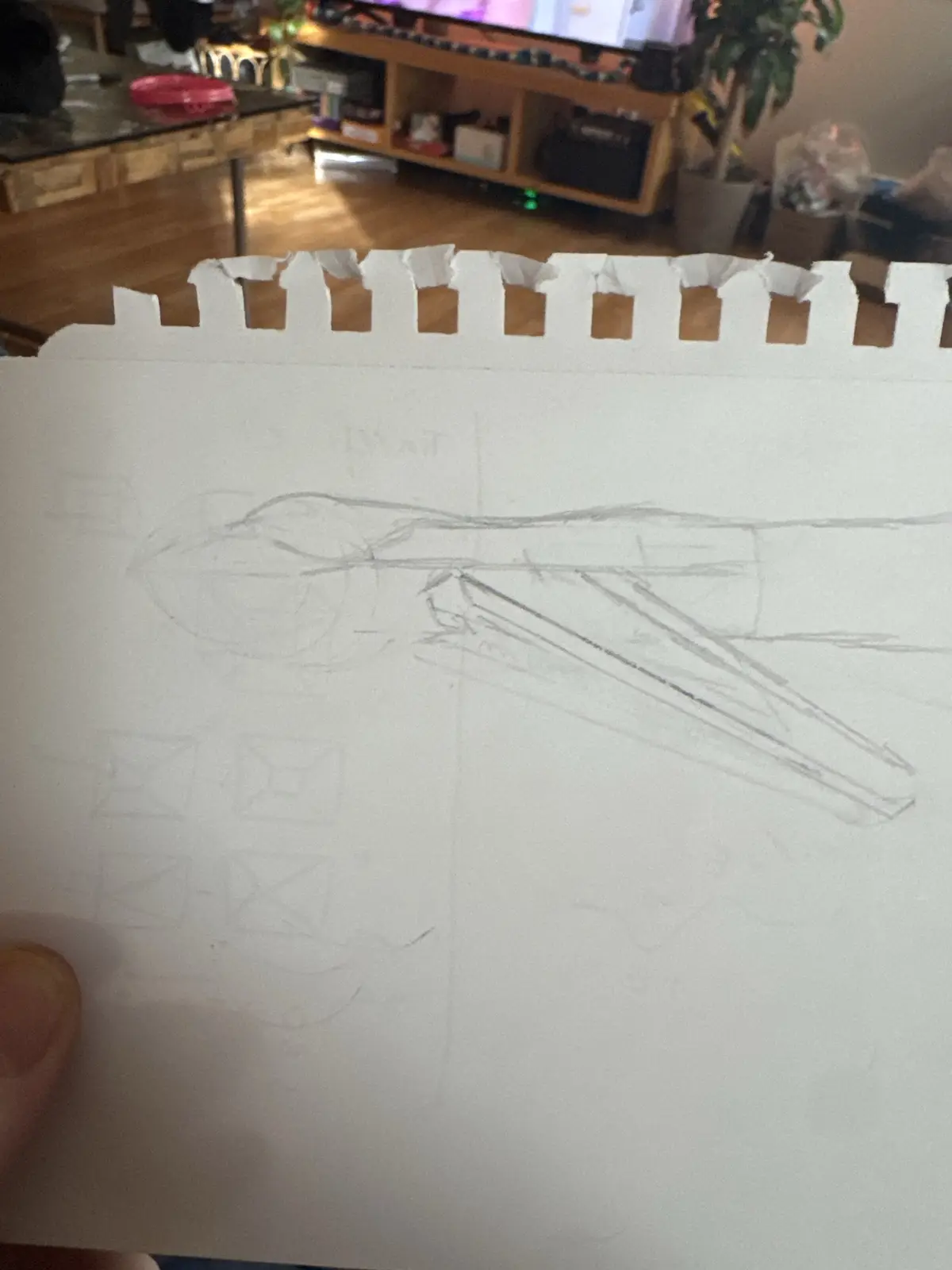 A hand holds up a piece of torn notebook paper with a rough pencil sketch of a long, angular object resembling a spaceship…