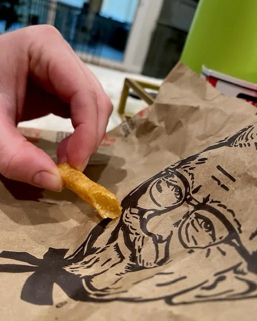 A close-up photo shows a person's hand holding a single, partially eaten french fry above a brown paper bag. The bag…