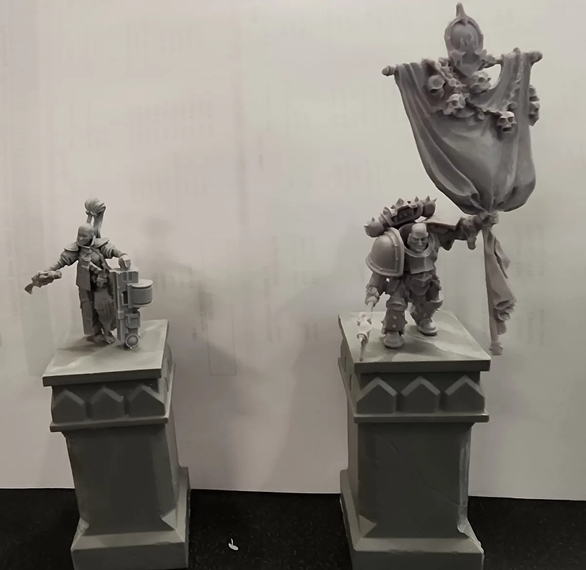 Two unpainted miniature figurines, one holding a large weapon and the other carrying a skull-adorned banner, stand on…