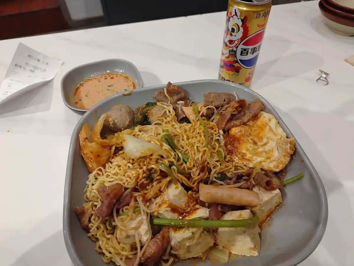 a close-up photo shows a large, grey rectangular bowl overflowing with ramen noodles mixed with various toppings including…