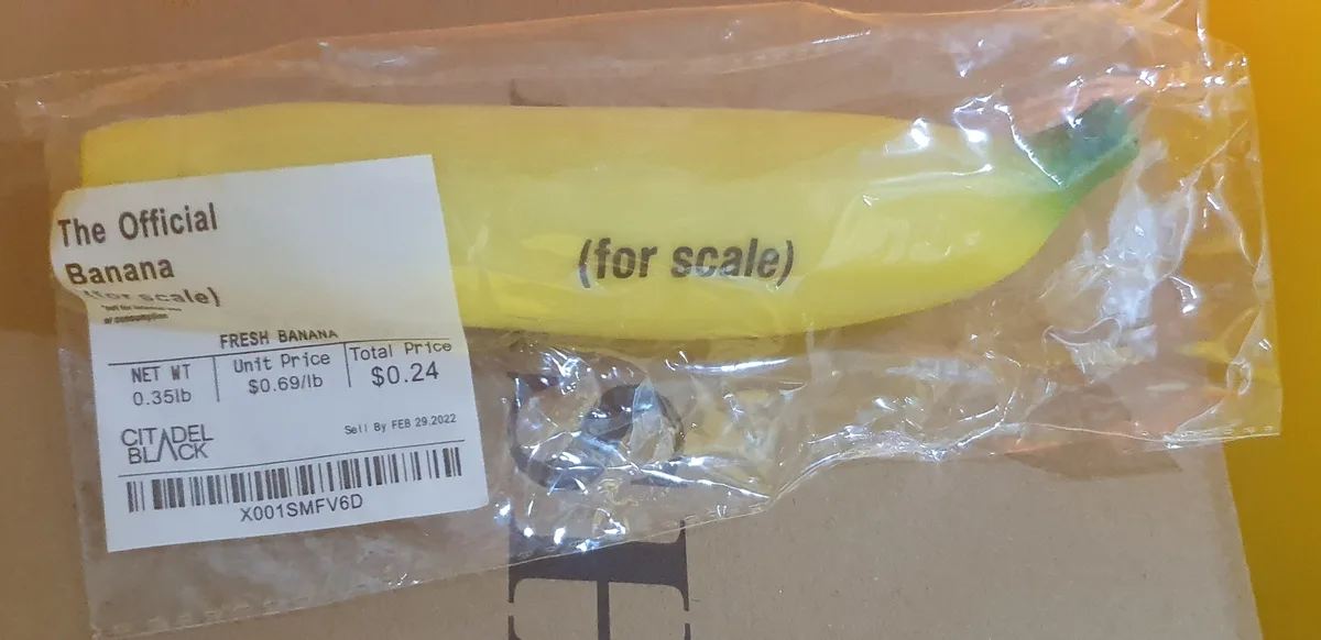 A yellow plastic banana-shaped object in clear plastic packaging, labeled as 'The Official Banana (for scale)' with a price…