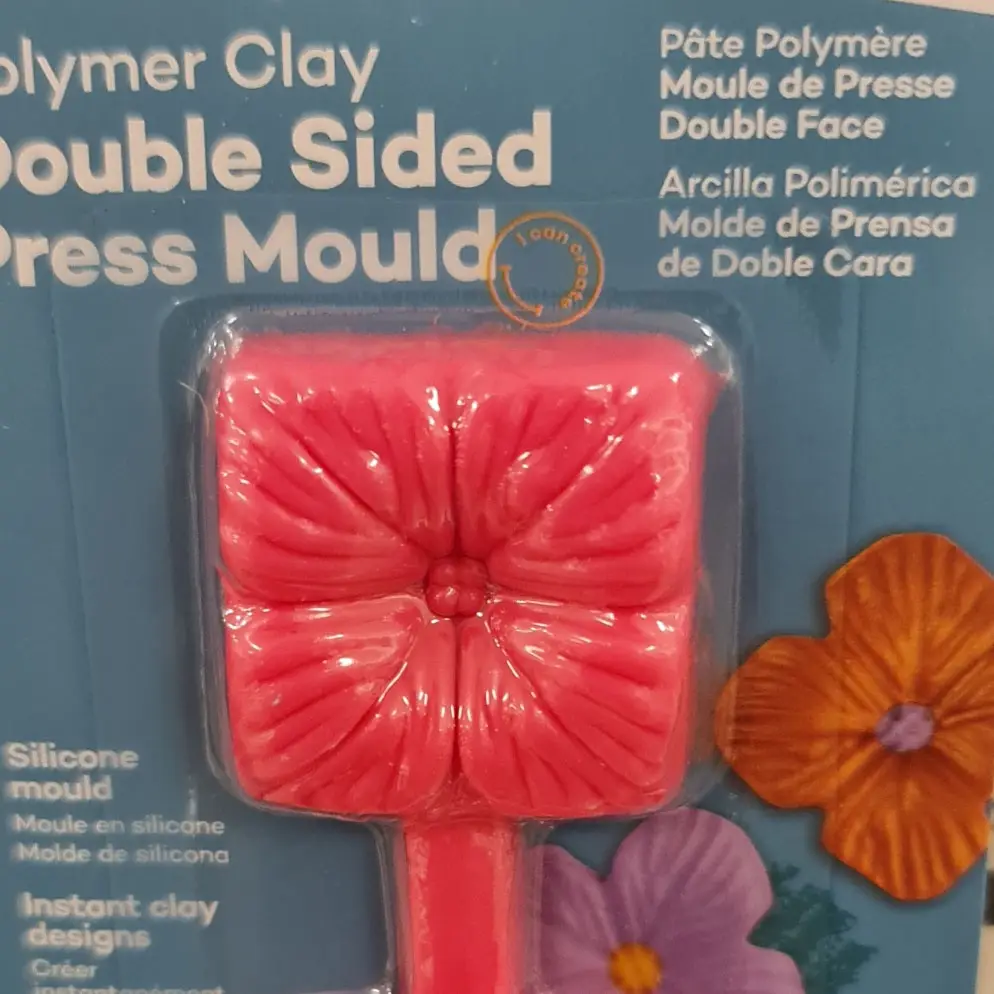 A close-up of a polymer clay press mould in its packaging, showing a pink, flower-shaped silicone mould with a square base…