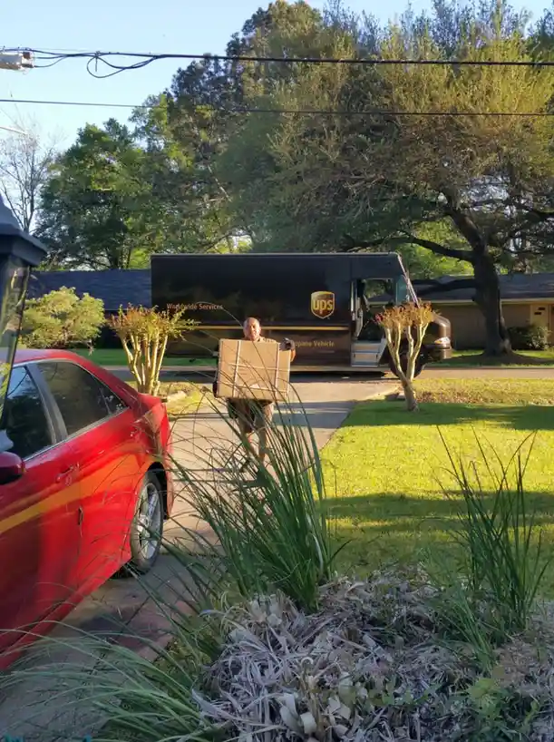 A UPS driver is carrying a large cardboard box down a residential sidewalk, with his black UPS truck visible behind him. A…