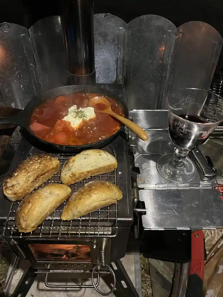 A cast-iron skillet with a steaming tomato-based stew topped with sour cream and herbs sits on a portable wood-burning…