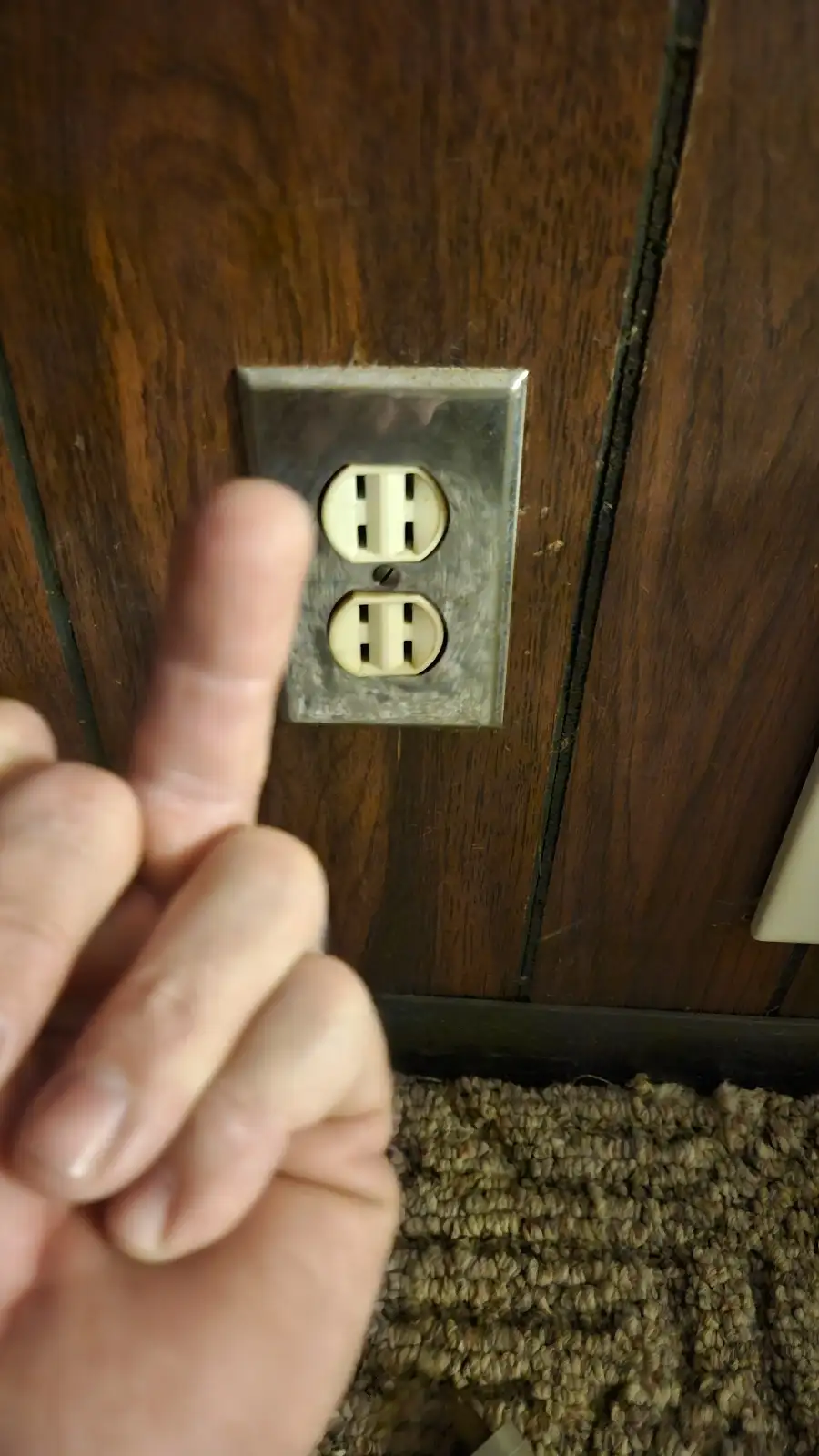A close-up photo shows a person's finger pointing directly at a standard North American electrical outlet mounted on a dark…