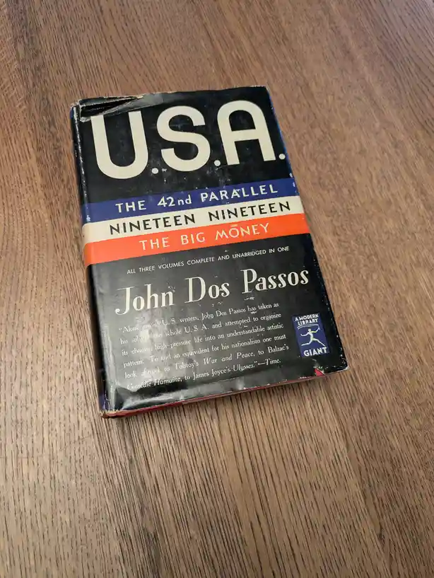 A well-worn paperback book titled 'USA' by John Dos Passos, featuring the three-part title 'The 42nd Parallel', 'Nineteen…
