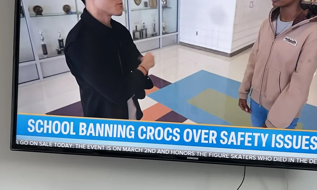 A television screen displays a news segment with two people standing in a room with colorful flooring and trophy cases in…