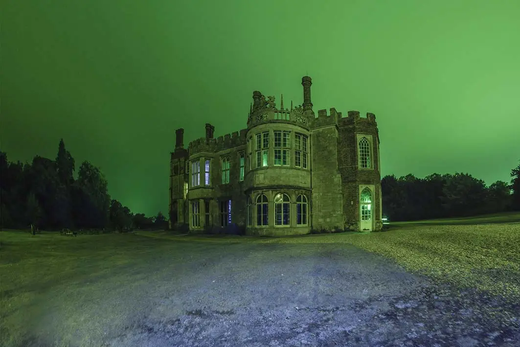 A large, old stone mansion stands at night under an unnaturally vibrant green sky, with some windows lit from within. The…
