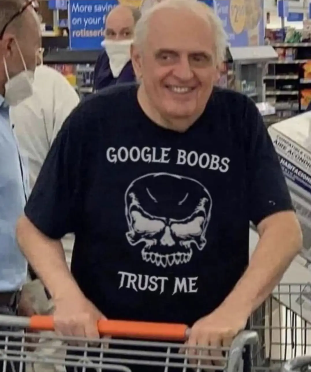 An older man with white hair is smiling broadly while pushing a shopping cart in a store. He wears a black t-shirt with a…
