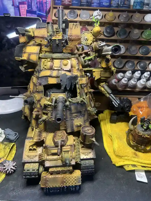 A heavily armed, yellow-painted Warhammer 40k tank model with multiple cannons and spikes sits on a workbench, surrounded by…