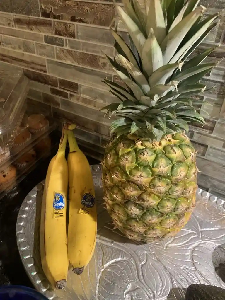 A large pineapple and two Chiquita bananas sit side by side on a decorative silver tray, with a tiled backsplash and a…