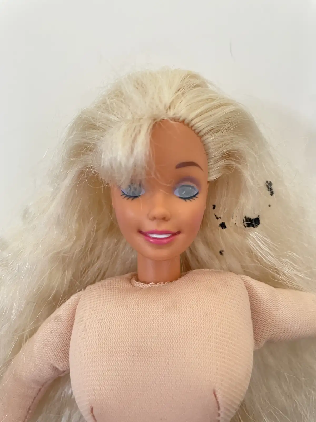 A close-up photo of a blonde Barbie doll with blue eyes and pink lips, wearing a beige knit sweater. The doll's hair has…