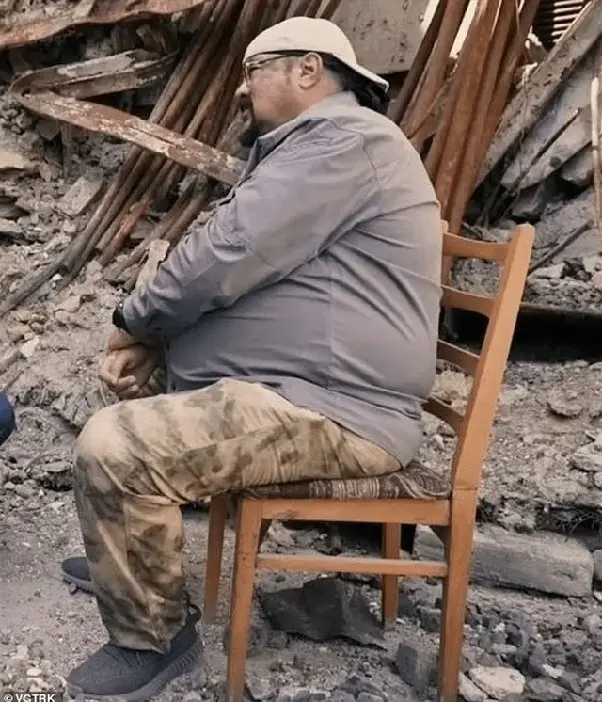 a man with a beard, glasses, and a cap sits calmly on a wooden chair amidst rubble and debris, wearing a grey shirt and…