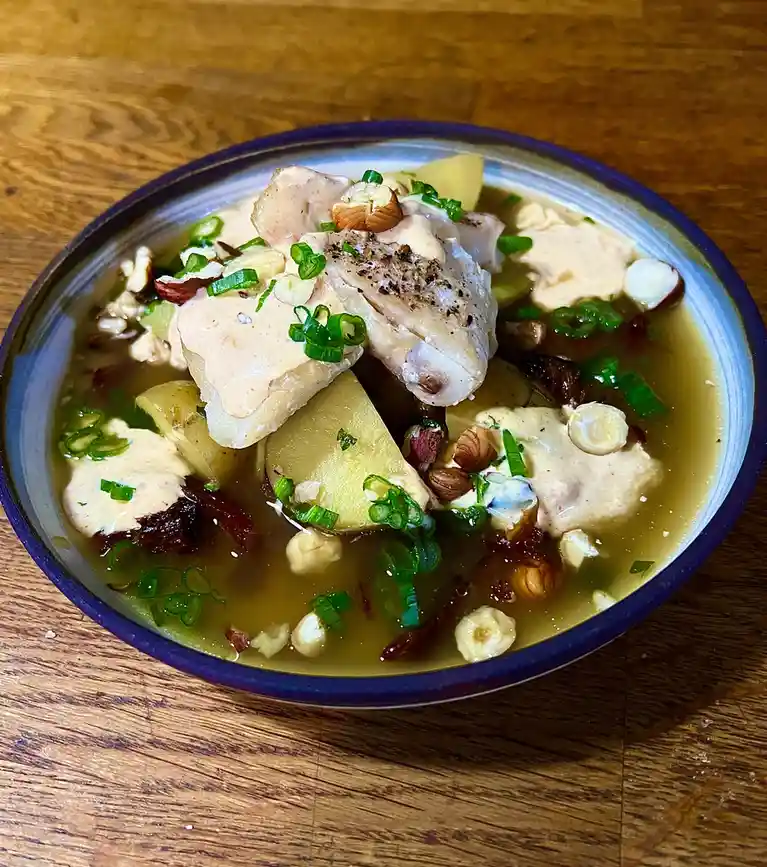 A close-up photo shows a bowl of soup on a wooden table, containing chunks of chicken, potato slices, chopped green onions…
