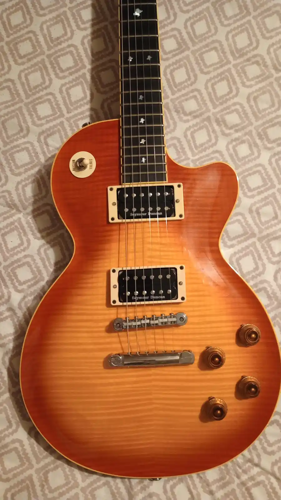 A close-up photo of a sunburst-finish electric guitar, likely a Gibson Les Paul style, resting on a patterned fabric…
