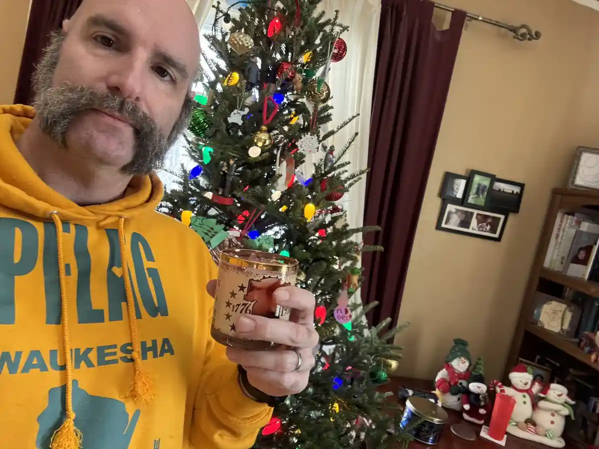 A man with a prominent mustache and goatee, wearing a yellow hoodie with 'PELAG Waukesha' printed on it, holds up a candle…