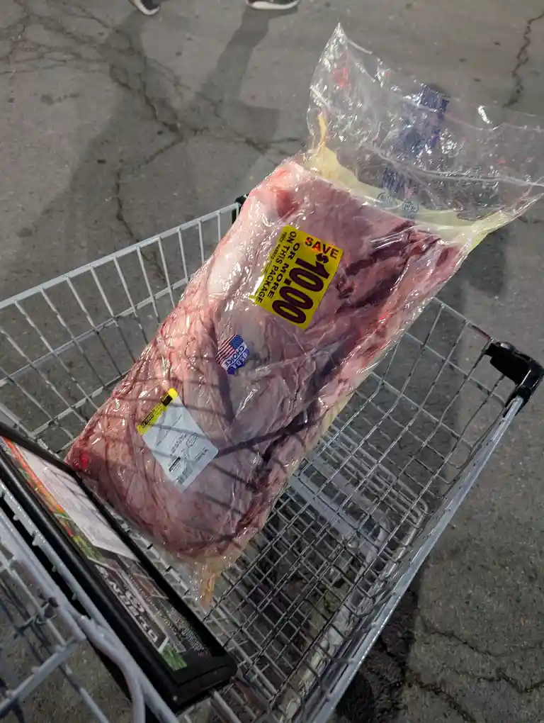 A large vacuum-sealed package of raw meat sits in a metal shopping cart, with a bright yellow sticker proclaiming 'SAVE…