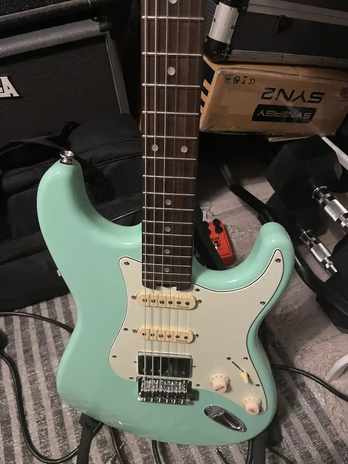A mint green electric guitar with a white pickguard and 'TESLA' branded pickups is shown upright, surrounded by music gear…