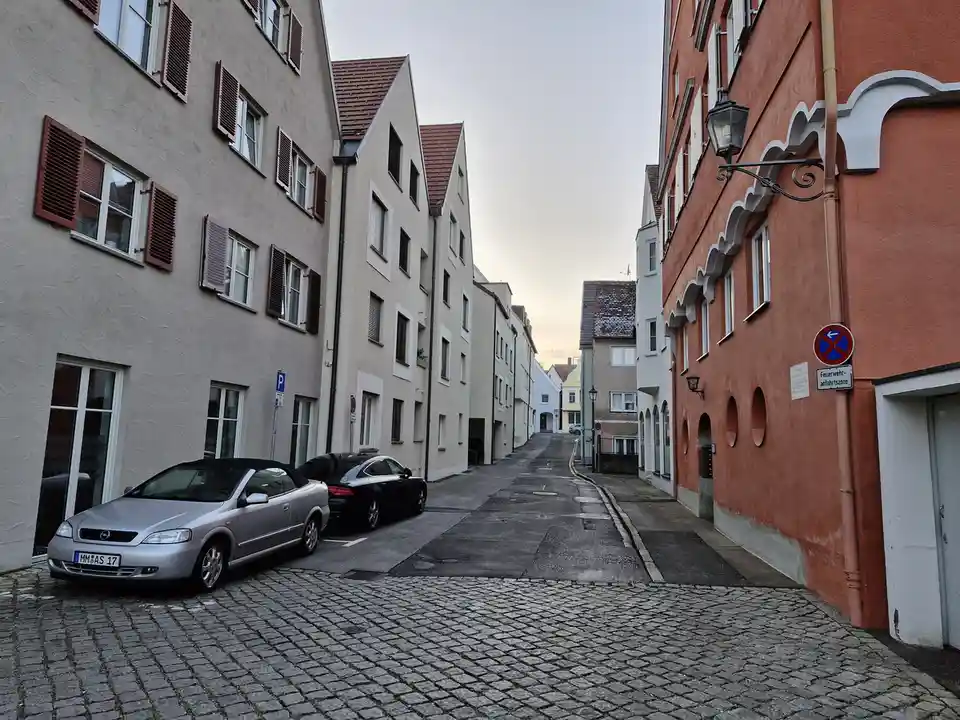 A narrow cobblestone street in a European town with parked cars and colorful buildings, featuring a blue and red circular…