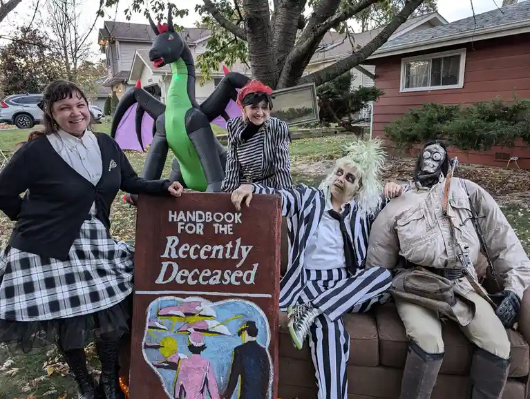 Four people in Halloween costumes pose outdoors with a large, hand-painted sign that reads 'HANDBOOK FOR THE Recently…