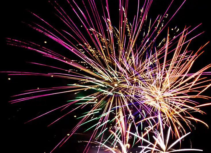 A vibrant nighttime photograph of colorful fireworks exploding in a dark sky, featuring streaks of magenta, gold, green, and…