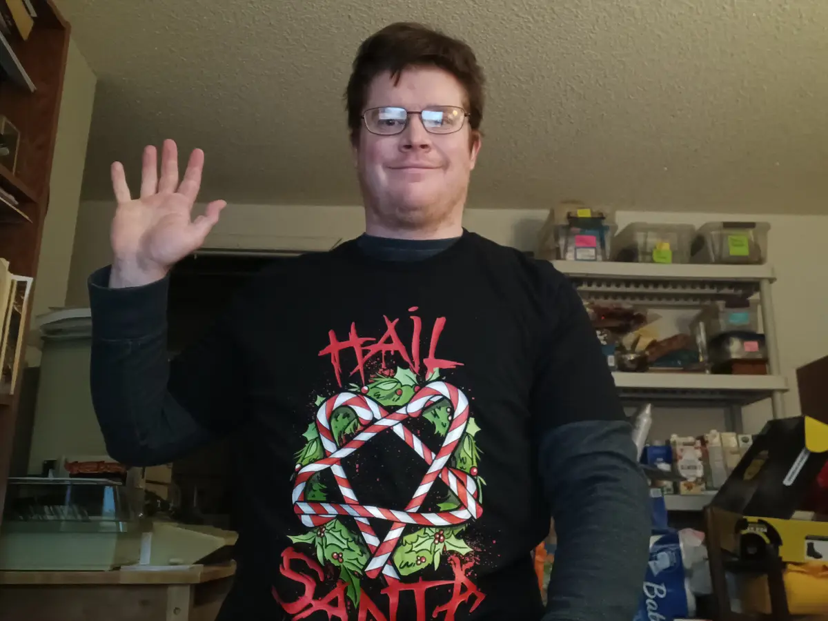 A man wearing a black holiday-themed t-shirt with 'HAIL SANTA' and candy cane graphics waves at the camera with a slight…