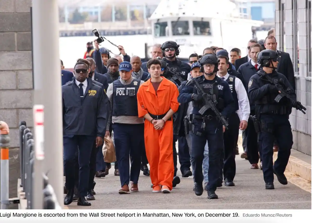 A man in an orange prison jumpsuit is being escorted by numerous NYPD officers, some in tactical gear with rifles, along a…