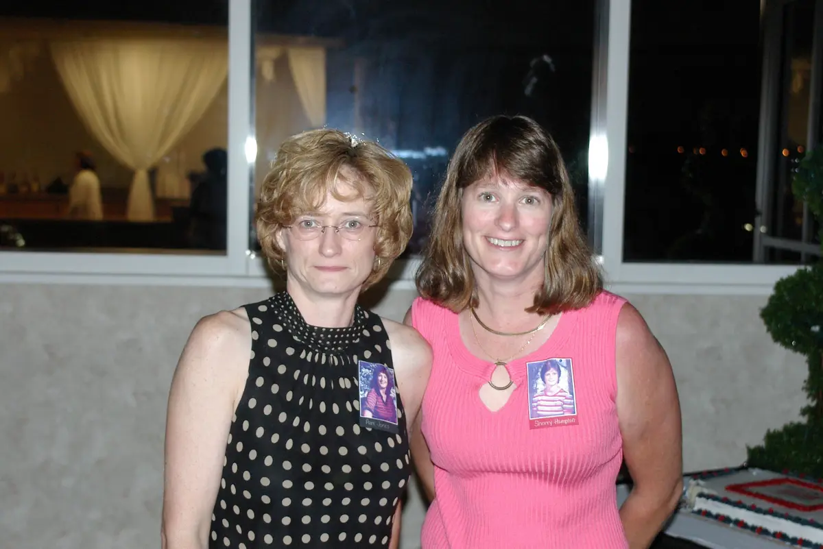 Two women pose for a photo at an event, both wearing name tags with photos of younger versions of themselves. The woman on…
