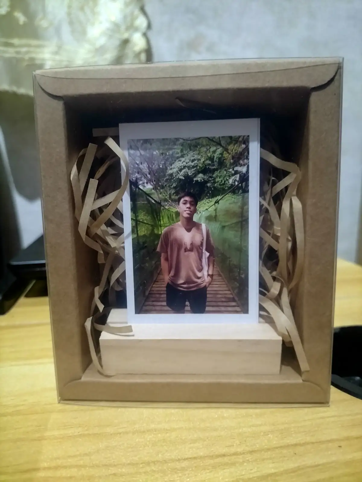 A photo of a young man standing on a wooden bridge surrounded by greenery is displayed inside a handmade cardboard box with…