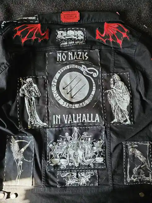 A black denim jacket covered in patches, including red bat wings, a central patch reading 'NO NAZIS IN VALHALLA' with a…