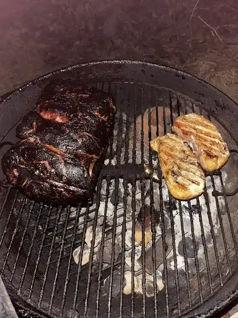 A close-up photo shows a round charcoal grill with a large, heavily charred piece of meat on the left and two smaller…