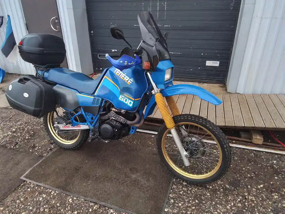 A blue Yamaha Ténéré 600 motorcycle with yellow front forks, black saddlebags, and a top case is parked on a mat outside a…