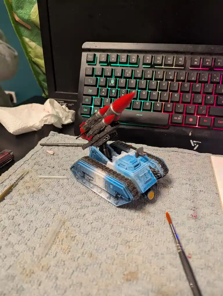 A painted miniature tank model with a red rocket launcher mounted on it sits on a grey towel in front of a black gaming…