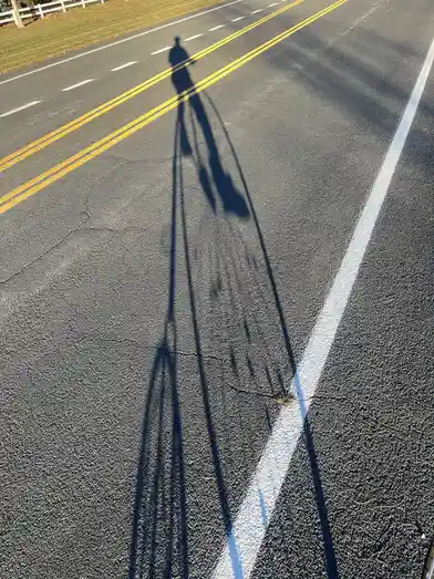A long shadow of a person standing on an asphalt road is cast by the low sun, stretching across the road's surface and…