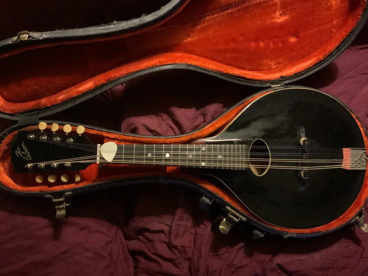 A black Gibson mandolin rests inside its open, red velvet-lined case, which sits on a dark red fabric surface. The…