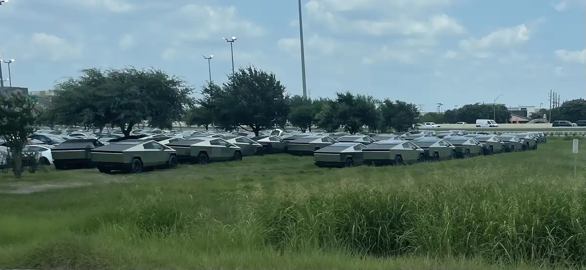 A wide shot shows dozens of silver Tesla Cybertruck vehicles parked in neat rows on a grassy field next to a paved parking…