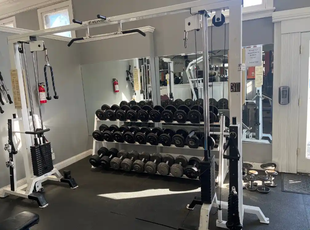 A well-lit gym interior showing a rack of neatly organized dumbbells, various weight machines, mirrors, and a fire…