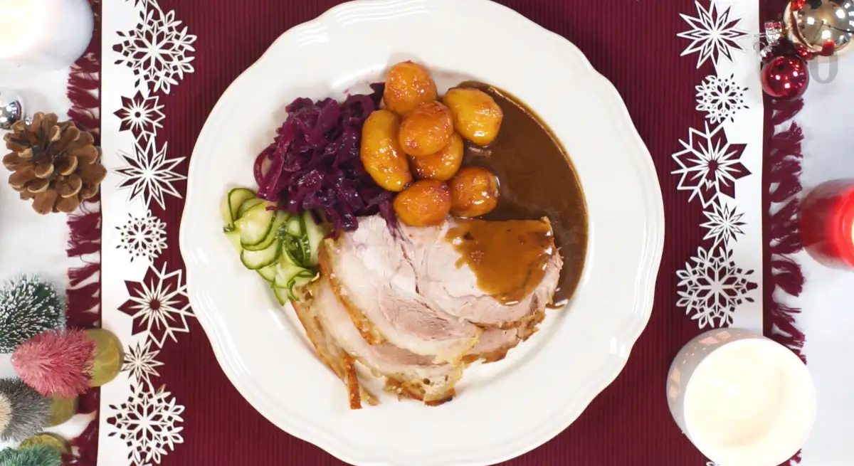 An overhead shot of a festive holiday table setting with a white plate holding sliced pork roast, red cabbage, glazed…