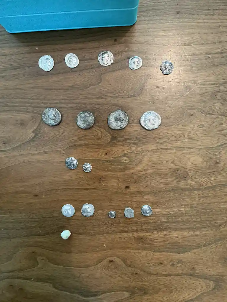 Several ancient Roman coins are arranged in rows on a wooden surface, with a small, light green, irregularly shaped stone at…