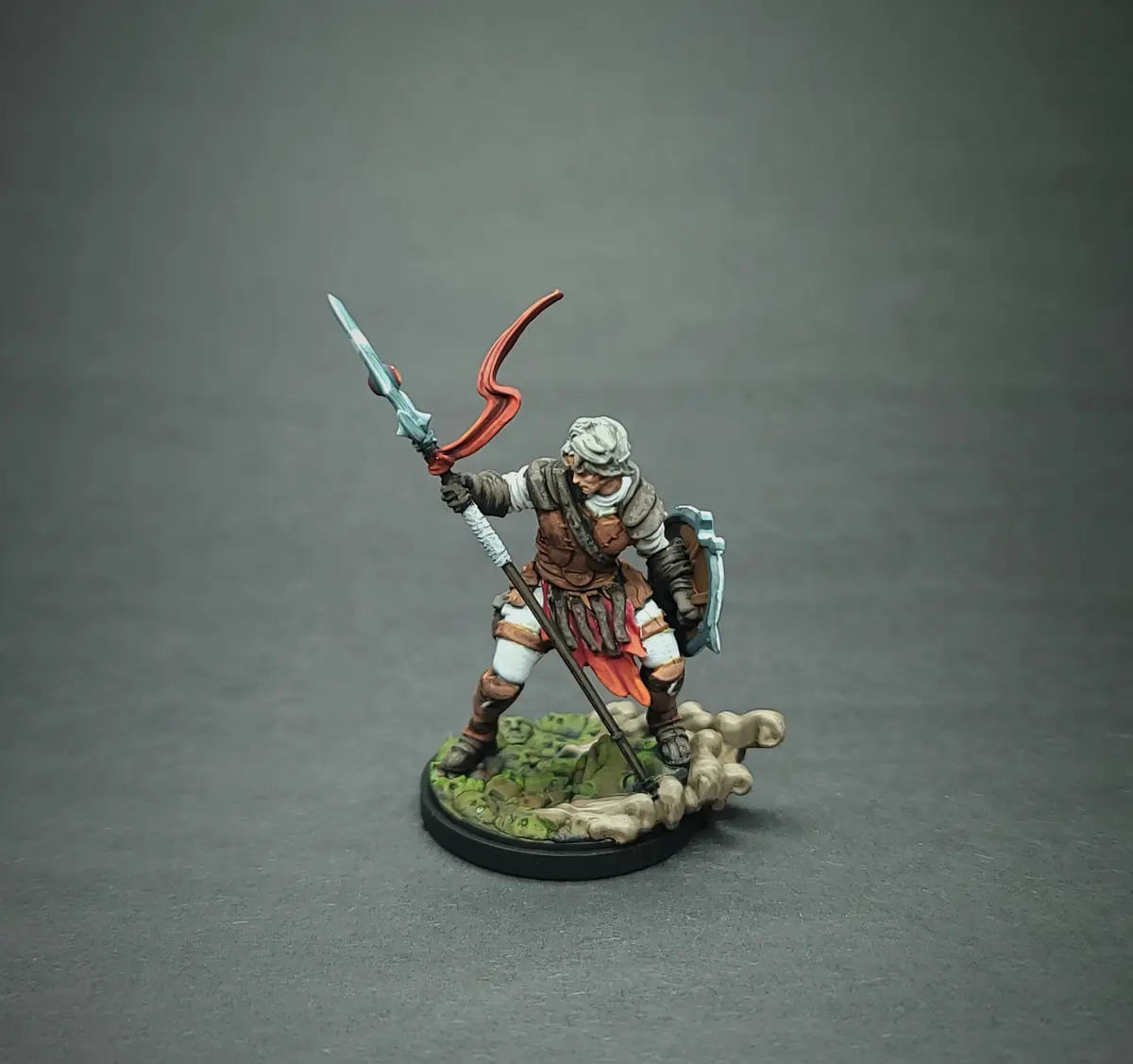 A painted miniature figurine of a warrior in a dynamic combat pose, holding a sword with a red curved blade and a shield on…