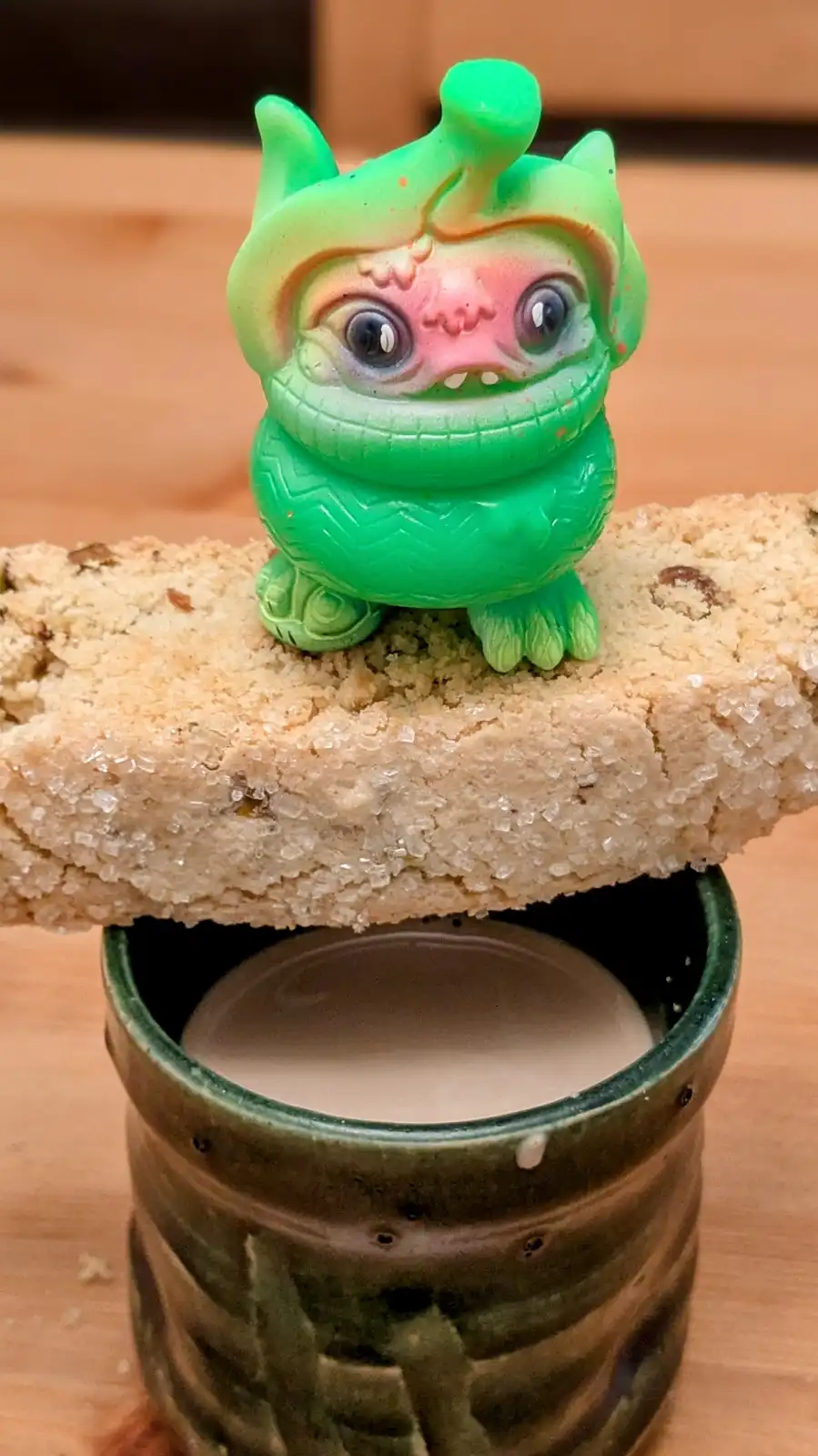 A small, green, cartoonish monster figurine with large eyes and a pink face sits atop a sugar-coated cookie that is balanced…