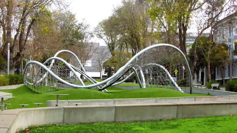 A large, modern, silver-colored metal playground structure with netting and curved tubes sits atop a grassy mound in a park…