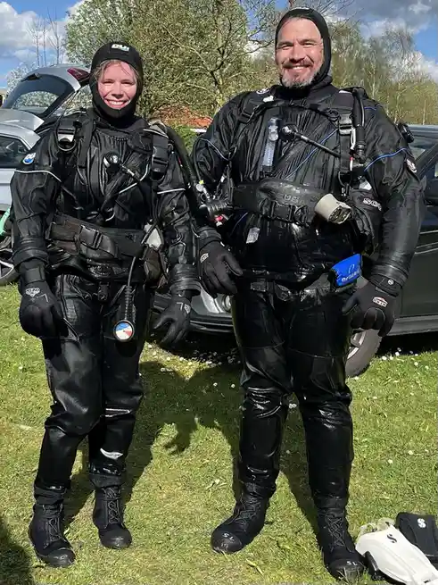 Two people in full scuba diving gear, including wetsuits, hoods, gloves, and oxygen tanks, stand smiling on a grassy field…