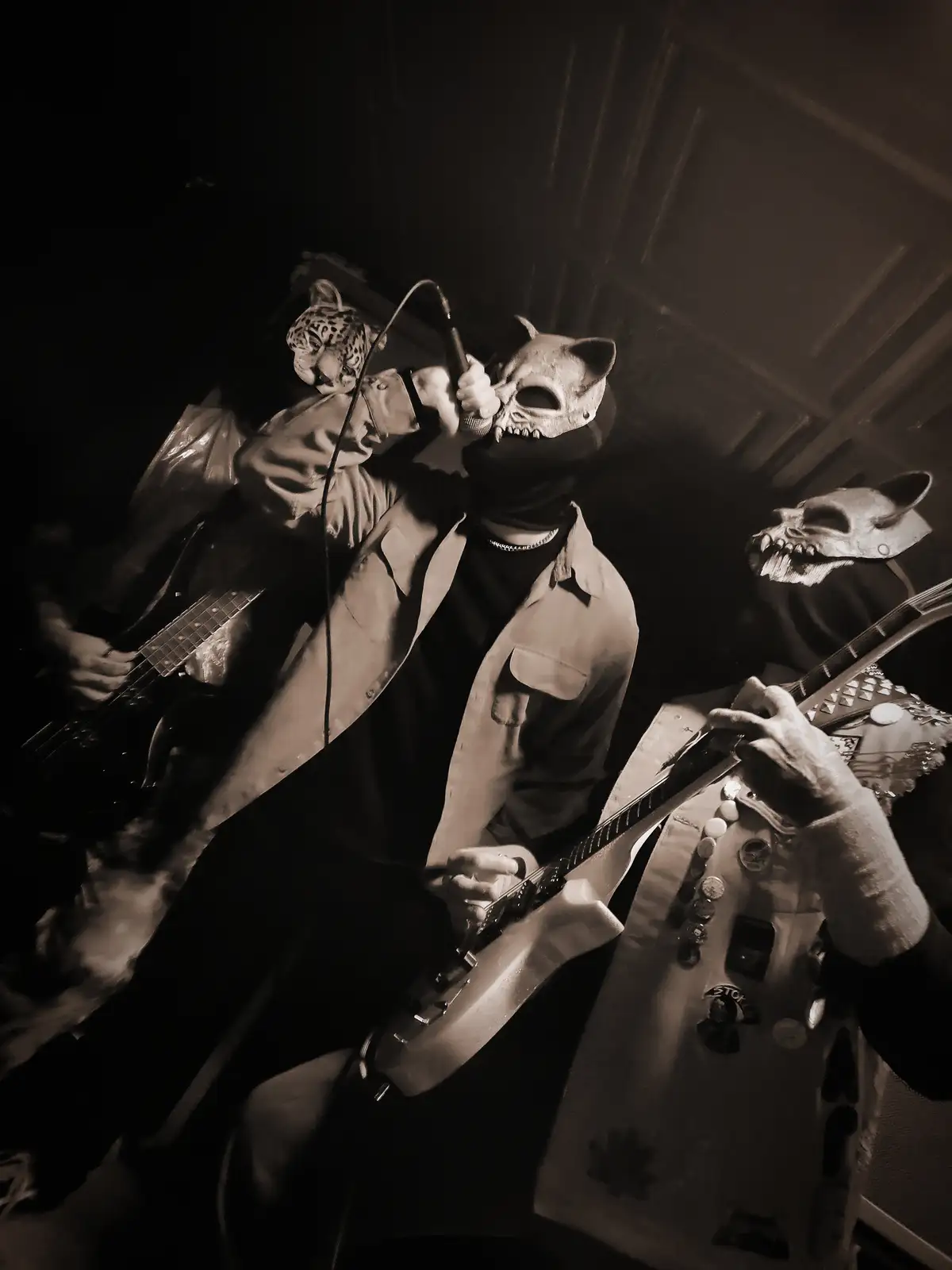A sepia-toned photo of a band performing on stage, with three musicians wearing animal masks — one leopard, one cat skull…