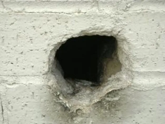 A close-up photo shows a rough, jagged hole punched through a light gray concrete block wall, revealing darkness inside. The…